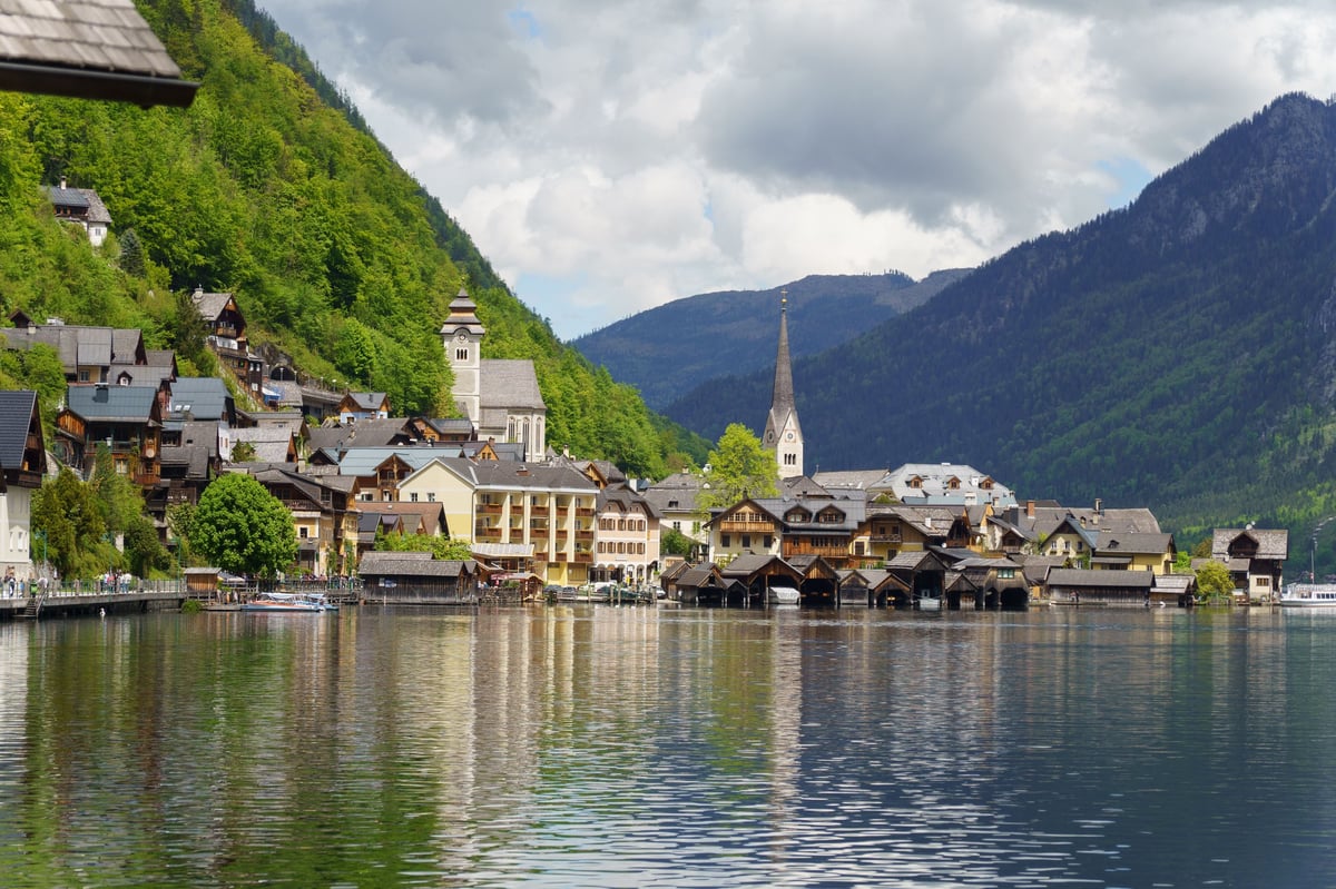 Alpenort mit Häusern am Wasser und Kirchturm vor steilen bewaldeten Bergen